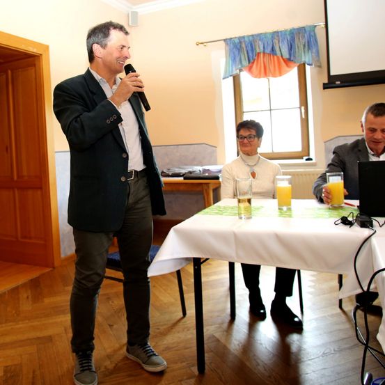 Ein Mann steht und spricht in ein Mikrofon, während ein anderer Mann und eine Frau an einem Tisch sitzen, mit Orangensaft vor ihnen.