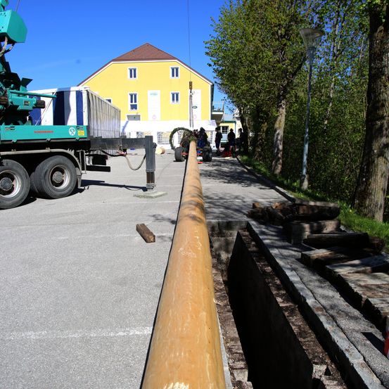 Ein Baufahrzeug steht auf einem Parkplatz, eine große Rohrleitung wird verlegt. In der Nähe stehen Menschen, möglicherweise bei der Arbeit. Ein gelbes Haus steht im Hintergrund.