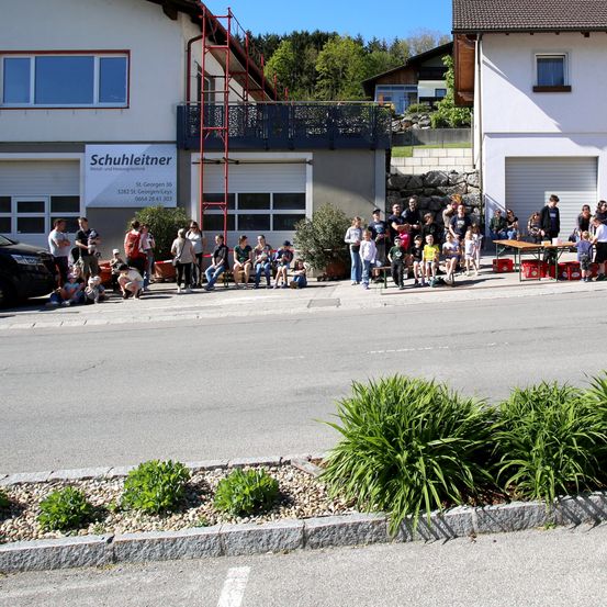 Eine Menschenmenge, einschließlich Kinder, versammelt sich vor einem Gebäude mit dem Schild 'Schuhleitner'. Einige sitzen auf der Bordsteinkante, andere stehen. Ein Tisch mit Gegenständen ist in der Nähe. Bäume und ein geparktes Auto sind im Hintergrund.