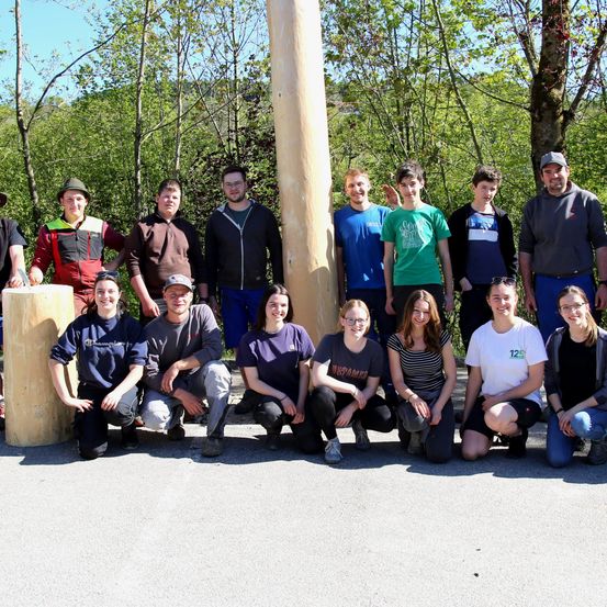 Eine Gruppe von Menschen posiert für ein Foto in einem Wald, einige knien und andere stehen. Sie tragen Freizeitkleidung, und Bäume umgeben sie. Ein Holzpfahl steht in der Mitte.