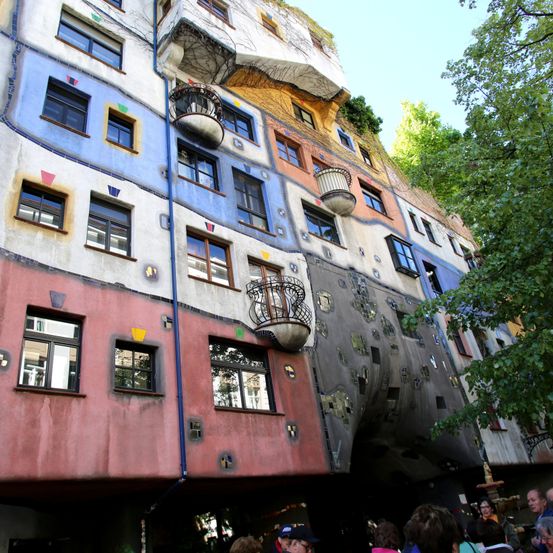 Ein mehrfarbiges Gebäude mit mehreren Balkonen und Fenstern steht unter einem klaren Himmel mit Bäumen. Menschen sind davor versammelt.
