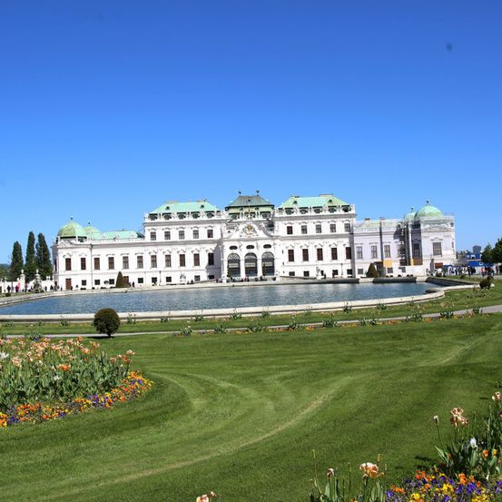 Ein großes weißes Gebäude mit grünen Kuppeln und einem Brunnen davor, umgeben von üppigen grünen Rasenflächen und leuchtenden Blumen unter einem klaren blauen Himmel.