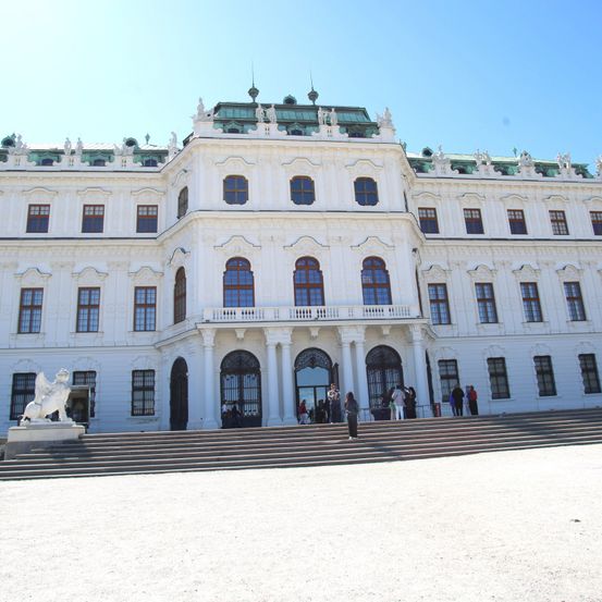 Ein großes weißes Gebäude mit grünem Dach, mehreren Fenstern und einer Löwenstatue davor. Menschen gehen die Stufen hinauf.