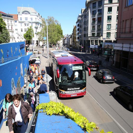 Ein roter Bus hält an einer Bushaltestelle. Menschen steigen ein. Mehrere Autos fahren auf der Straße. Gebäude säumen beide Straßenseiten.