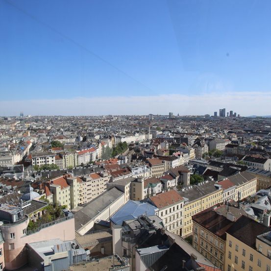 Ein Panoramablick von oben auf eine Stadt mit zahlreichen Gebäuden, einige mit roten Dächern, unter einem blauen Himmel mit Wolken.