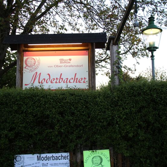 Ein Schild für Moderbacher ist in der Dämmerung beleuchtet. Dahinter steht eine Hecke mit einem Laternenpfosten. Der Himmel ist klar und blau.