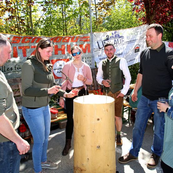 Menschen versammeln sich um einen Holzpfahl mit Nägeln, zwei halten Hämmer. Sie scheinen an einer Outdoor-Veranstaltung teilzunehmen. Bäume und ein Banner sind im Hintergrund.