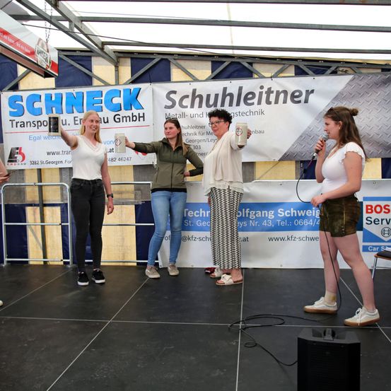 Vier Frauen stehen auf der Bühne, halten Tassen und lächeln. Ein Banner mit dem Text 'Schuhleitner' hängt hinter ihnen. Zwei Frauen halten Mikrofone. Die Bühne hat einen schwarzen Boden.