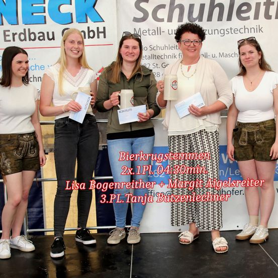 Fünf Frauen stehen auf einer Bühne und lächeln. Sie halten Auszeichnungen und Bierkrüge. Die Frau in der Mitte trägt eine Brille und hält einen Papierpreis. Der Hintergrund hat ein Banner mit weißem Text.