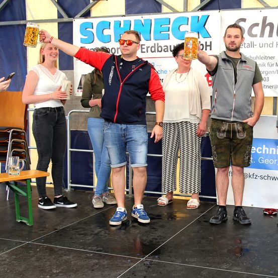 Vier Personen stehen auf der Bühne eines Bierfestivals, wobei zwei Männer Biergläser halten. Im Hintergrund steht ein Schild mit der Aufschrift SCHNECK.