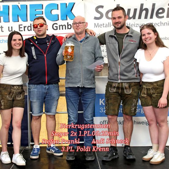 Fünf Personen stehen lächelnd auf einer Bühne und halten ein Bier. Zwei Männer tragen traditionelle bayerische Shorts. Der Mann in der Mitte hält ein Stück Papier. Ein Banner hinter ihnen steht 'Schneider.'