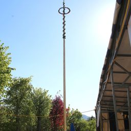 Ein hoher Holzpfahl mit einer Spiralgirlande steht in einem Park mit Bäumen. Ein Gebäude mit Metallrahmen befindet sich daneben.