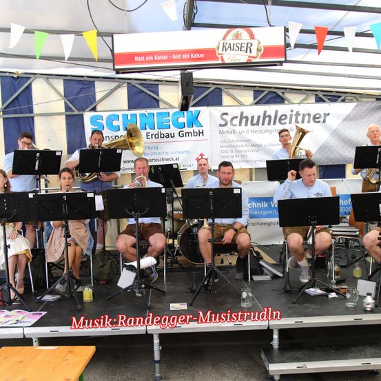Eine Band spielt auf der Bühne. Mehrere Personen sitzen auf Stühlen und spielen verschiedene Instrumente, darunter Trompeten, Posaunen und Trommeln. Ein Banner mit dem Wort 'Kaiser' ist über der Bühne angebracht.