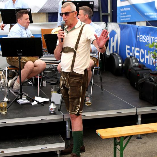 Ein Mann in traditioneller bayerischer Tracht spricht in ein Mikrofon auf der Bühne, während eine Band hinter ihm spielt. Die Bühne ist mit verschiedenen Musikinstrumenten und Ausrüstung ausgestattet.