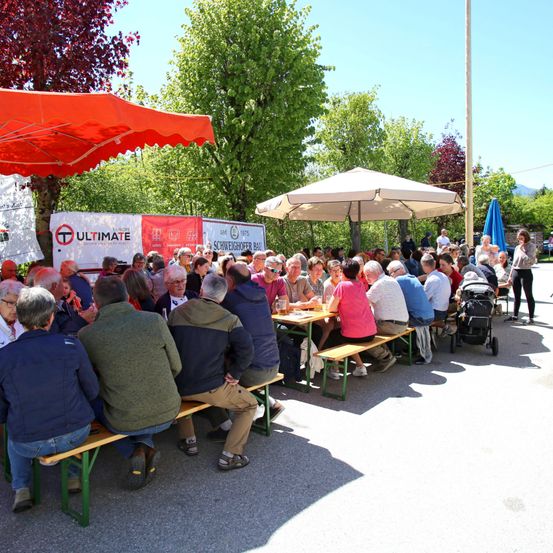 Viele Menschen sitzen um Tische unter Schirmen. Die Umgebung ist im Freien mit Bäumen und Bergen im Hintergrund. Ein Kinderwagen ist in der Nähe zu sehen.