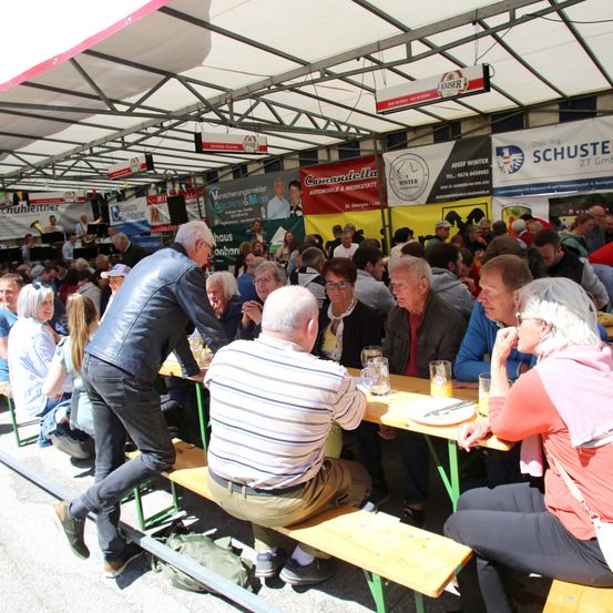 Eine Gruppe älterer Personen sitzt an einem großen Tisch im Freien, möglicherweise auf einem Festival. Sie unterhalten sich, einige essen, andere trinken. Über ihnen hängen Banner.
