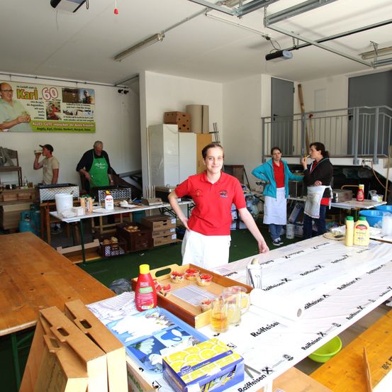 Ein Raum mit Leuten in Schürzen, die Essen zubereiten. Ein Mann in einem roten Hemd steht vor einem langen Tisch. Ein Banner hängt an der Wand.