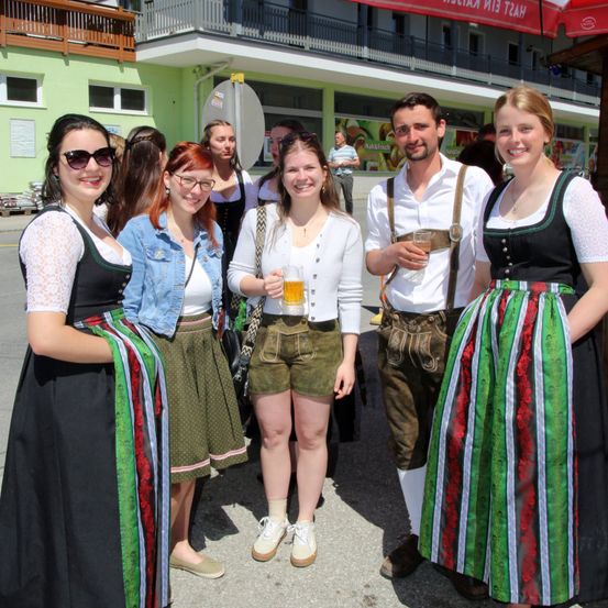 Eine Gruppe von Menschen in traditioneller deutscher Kleidung steht lächelnd vor einem grünen Gebäude, für die Kamera.