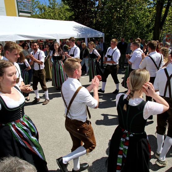 Eine Gruppe von Menschen in traditioneller deutscher Kleidung tanzt unter einem weißen Zelt, während Zuschauer im Hintergrund zusehen.