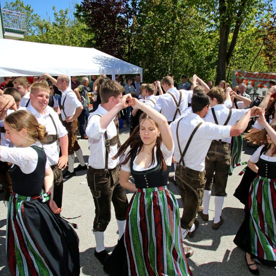 Eine Gruppe von Menschen in traditioneller bayerischer Kleidung tanzt im Freien mit Bäumen und einem Zelt im Hintergrund.