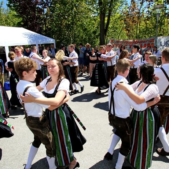 Menschen in traditioneller Kleidung tanzen im Freien im Kreis. Einige tragen Hosenträger und andere haben Blumen im Haar. Dahinter steht ein weißes Zelt und ein Banner.