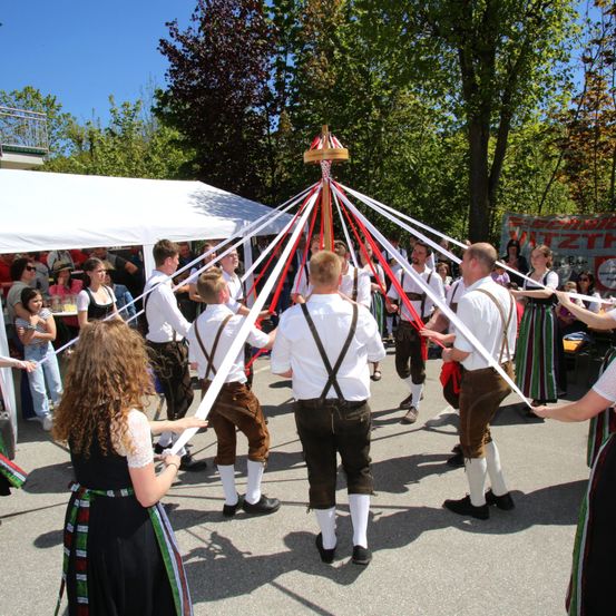 Eine Gruppe von Menschen in traditioneller Kleidung tanzt um einen Maibaum. Zuschauer beobachten hinter den Seilen. Ein Zelt und Bäume sind im Hintergrund.