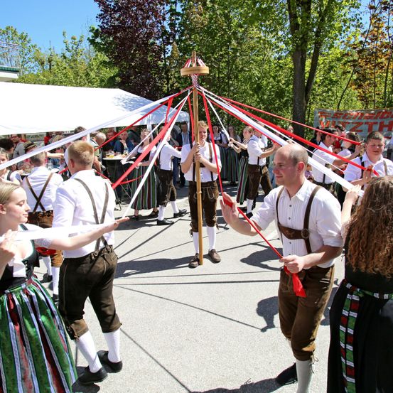Eine Gruppe von Menschen in traditioneller bayerischer Kleidung tanzt um einen Maibaum im Freien.