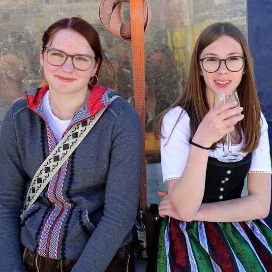 Zwei junge Frauen posieren für ein Foto. Die Frau auf der linken Seite trägt eine graue Jacke und eine Brille. Die Frau auf der rechten Seite hält ein Weinglas und trägt ein traditionelles Kleid.