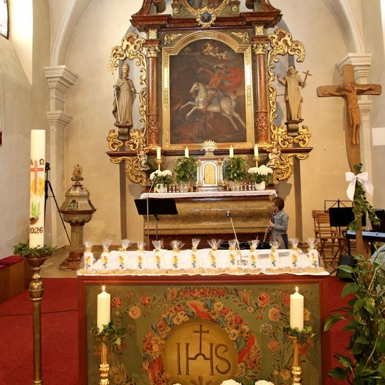 Ein Altar in einer Kirche mit einem Gemälde, Statuen und Kerzen. Eine junge Person spielt Klarinette. Es gibt Pflanzen und Blumen in der Umgebung.