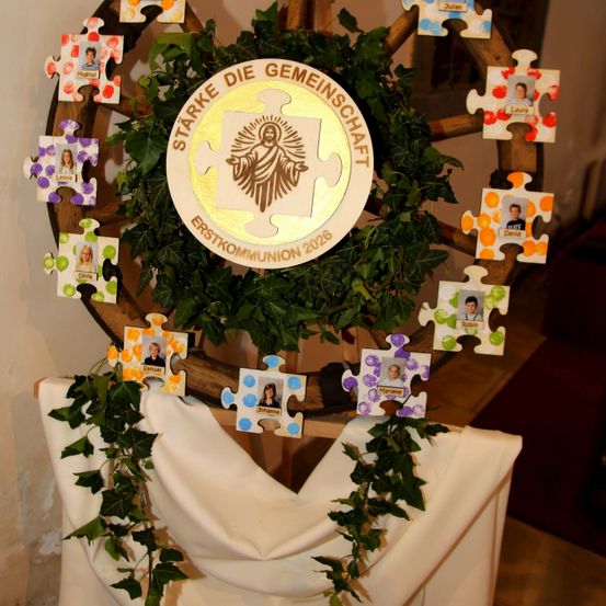 Eine große runde Anzeige mit einem zentralen Emblem, umgeben von Puzzleteilen mit Fotos von Kindern. Das Emblem trägt die Aufschrift 'Stärke die Gemeinschaft' und 'Erstkommunion 2026'. Grüner Efeu ist um die Anzeige gelegt.