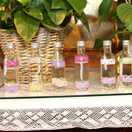 Sechs kleine klare Flaschen mit Wasser, jede mit anderen Namen und rosa Spitzendesigns beschriftet. Hinter den Flaschen hält ein Korb eine grüne Pflanze.