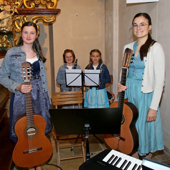 Vier Mädchen posieren mit Musikinstrumenten in einer Kirche. Zwei halten Gitarren, eine spielt Klarinette und eine andere Flöte. Sie stehen neben einem Klavier und Notenständern.