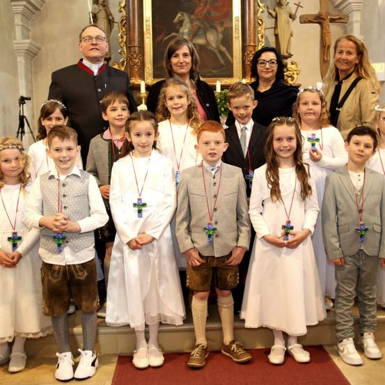 Eine Gruppe von Kindern in weißen Kleidern mit bunten Kreuzen um den Hals posiert mit Erwachsenen in einer Kirche für ein Foto.