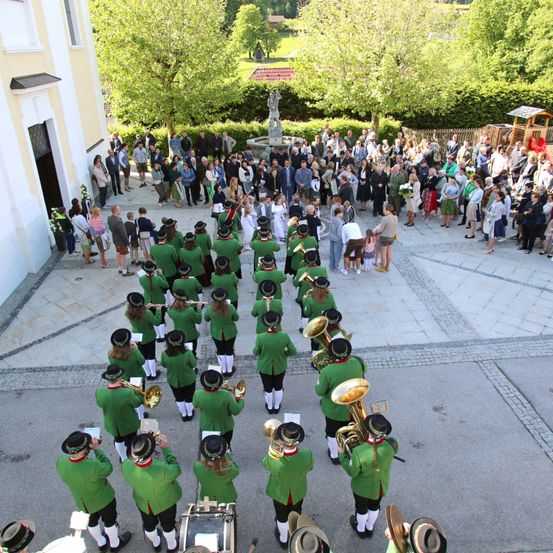 Eine große Menschenmenge versammelt sich vor einem Gebäude mit einer Statue. Eine Blaskapelle in grünen Uniformen tritt auf dem gepflasterten Platz auf. Viele Menschen beobachten die Aufführung.