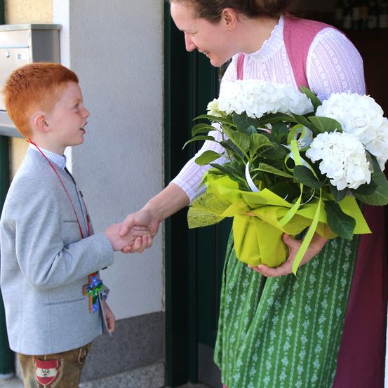 Eine Frau in einem grünen Kleid, die einen weißen Blumenstrauß hält, schüttelt einem jungen Jungen in einem grauen Jackett die Hand.