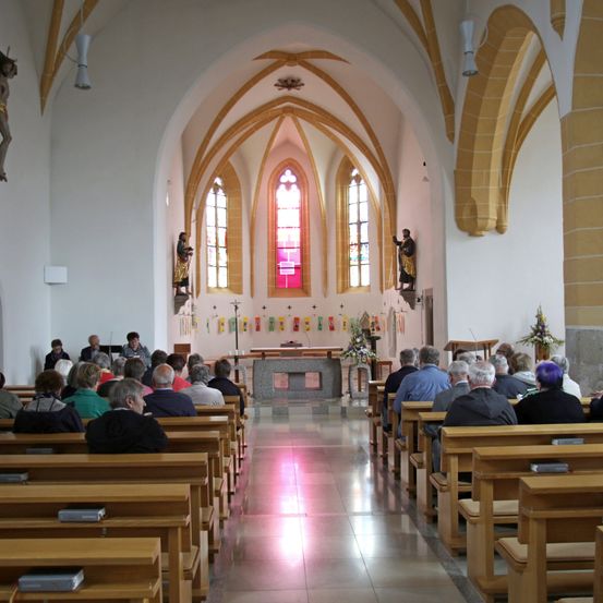 Ein Innenraum einer Kirche mit Holzbänken, auf denen Menschen sitzen und der Predigt lauschen. Der Altar ist vorne mit Blumen und Statuen an der Wand.