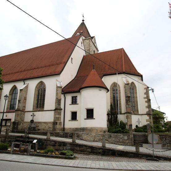 Eine weiße Kirche mit braunem Dach und Turm. Es gibt einige Fenster, Pflanzen und ein Kreuz davor. Ein Laternenpfosten befindet sich auf der linken Seite.