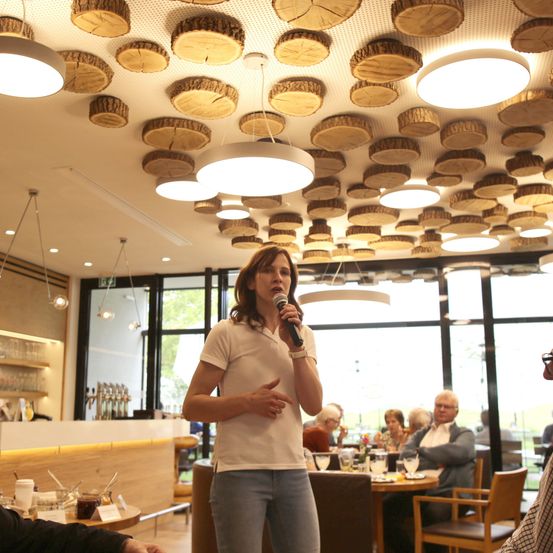 Eine Frau spricht in ein Mikrofon in einem Restaurant mit einer Holzscheit-Decke, umgeben von sitzenden Gästen. Der Raum hat Glaswände und eine Holzbar.