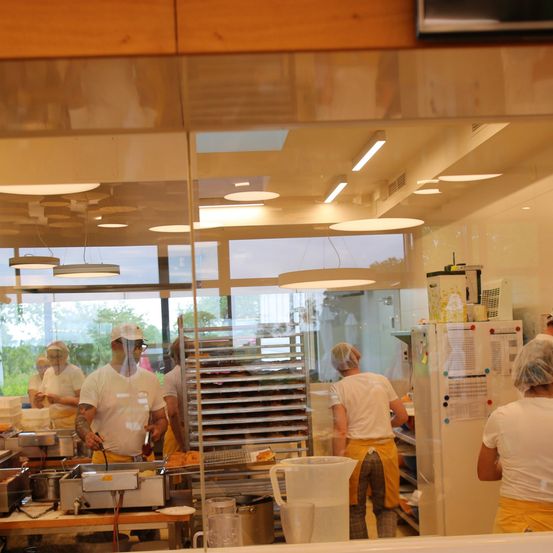 Ein Bäckerei-Interieur mit Arbeitern in weißen Uniformen und Haarnetzen. Einer kocht, während andere Teig zubereiten. Eine Glaswand reflektiert das Außen.