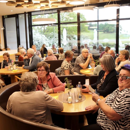 Eine Gruppe älterer Menschen sitzt in einem Restaurant. Sie unterhalten sich und einige halten Tassen. Auf dem Tisch stehen eine Vase, eine Flasche und ein Glas. Das Restaurant hat Glaswände und -türen.