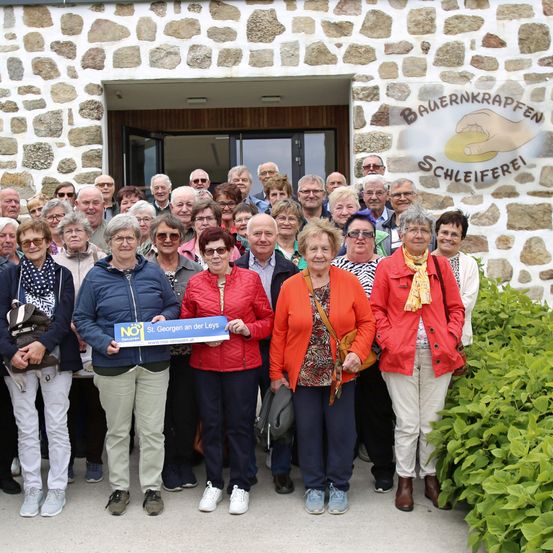 Eine Gruppe älterer Menschen posiert für ein Foto vor einem Gebäude mit einer Steinmauer. Das Gebäude hat ein Schild mit der Aufschrift "BAUENKRAPFEN SCHLEIFREI".