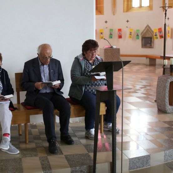 Drei Personen sitzen auf einer Bank in einer Kirche, jede hält ein Buch. Ein Mikrofon ist vor ihnen auf einem Podest aufgestellt.