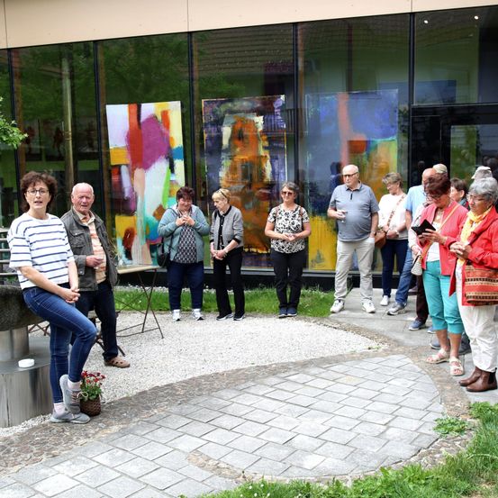 Eine Gruppe von Menschen versammelt sich vor einer Kunstgalerie, einige schauen sich die Gemälde an. Es gibt abstrakte Gemälde an den Glaswänden. Eine Frau vorne hält ein Buch und scheint es zu lesen.