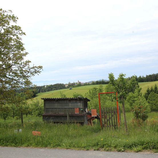 Ein Schuppen mit einem roten Tor steht in einem Grasfeld mit einem entfernten Hügel. Bäume umgeben das Gebiet, und es ist ein kleiner Obstgarten zu sehen.