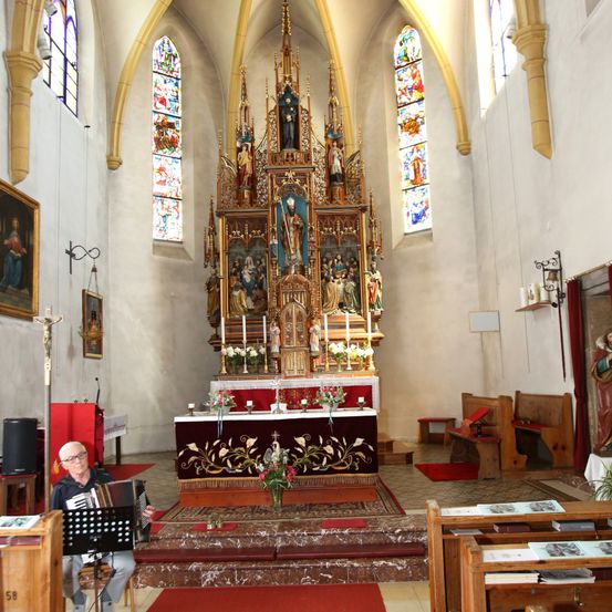 Ein Mann spielt Akkordeon in einer Kirche. Der Altar hat ein aufwendiges Design. Buntglasfenster und Statuen schmücken die Wände.