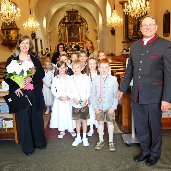 Eine Gruppe von Kindern und Erwachsenen posiert in einer Kirche, mit einem Priester und einer Frau im Vordergrund. Die Kinder tragen traditionelle bayerische Kleidung.