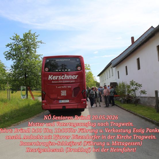 Ein roter Bus mit Kerschhner Reisen darauf steht auf einer Straße. Leute stehen draußen. Ein Gebäude mit einem Schornstein ist rechts. Bäume und Gras sind links.