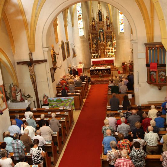 Ein Kircheninneres mit Menschen, die in hölzernen Kirchenbänken sitzen, ein Priester steht am Altar mit einem roten Teppich, der dorthin führt. Ein Kreuz und eine Statue schmücken die Wände.