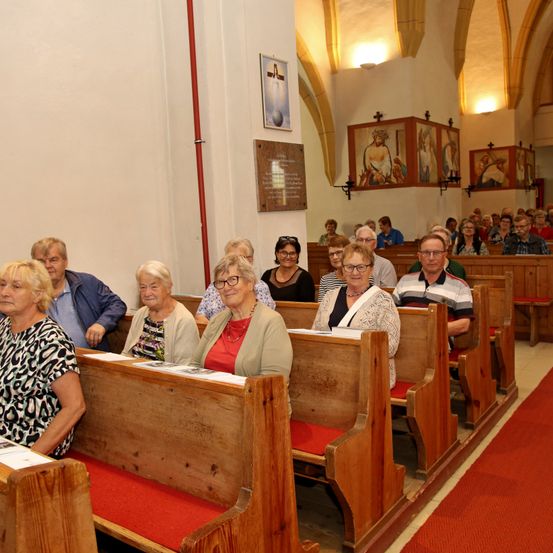Mehrere Menschen sitzen in hölzernen Kirchenbänken, die nach vorne gerichtet sind. Einige tragen Brillen und einer hält ein Papier. Kunstwerke und ein Kreuz schmücken die Wand.