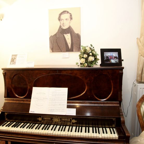 Ein antikes Klavier mit Notenpapier darauf, einem Blumenstrauß und einem Bilderrahmen mit einem Foto an der Wand.